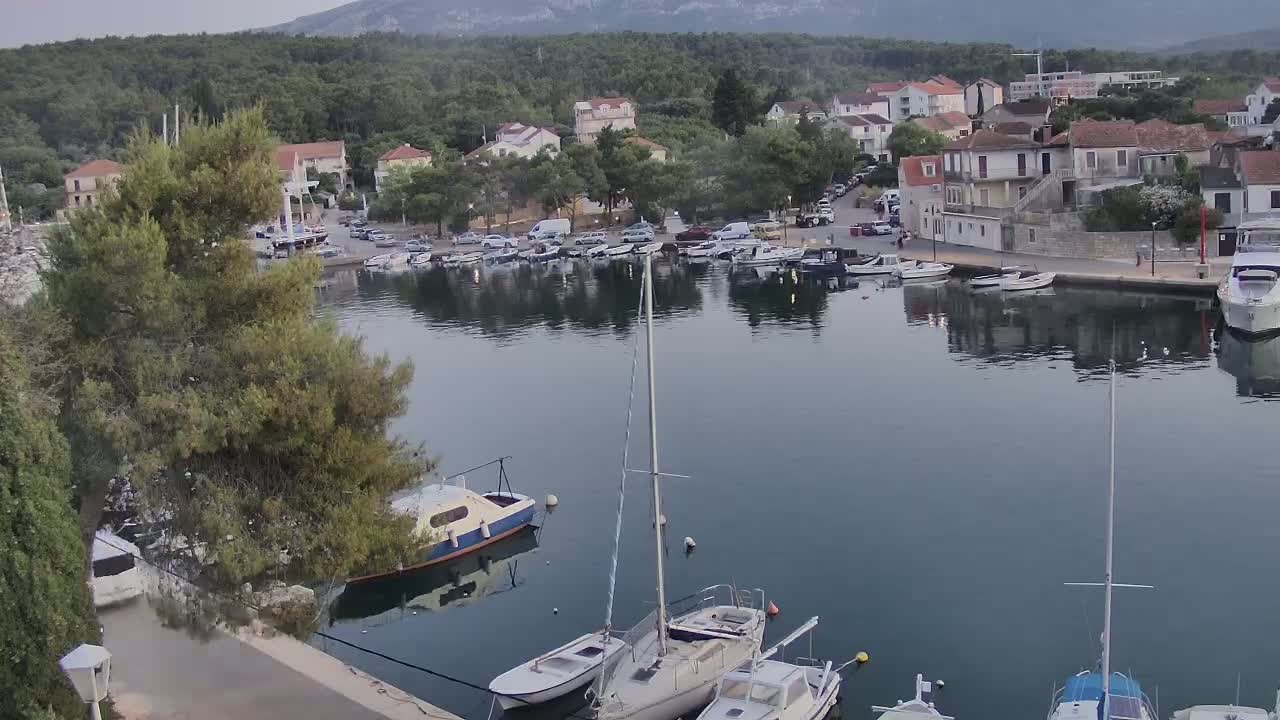 A calm harbor filled with sailboats and motorboats is nestled next to a village of low, light-colored buildings against a backdrop of green hills under a mostly cloudy sky.