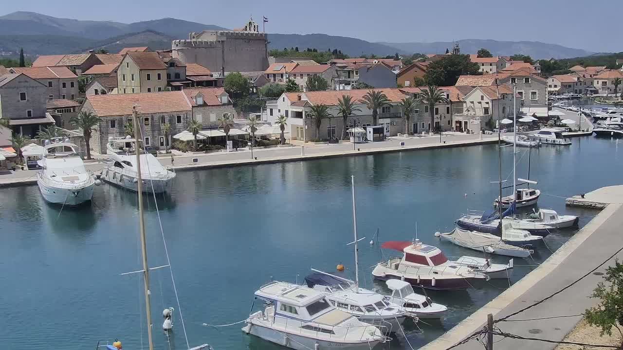 A harbor filled with boats is situated in front of a town with stone buildings and palm trees under a clear sunny sky.