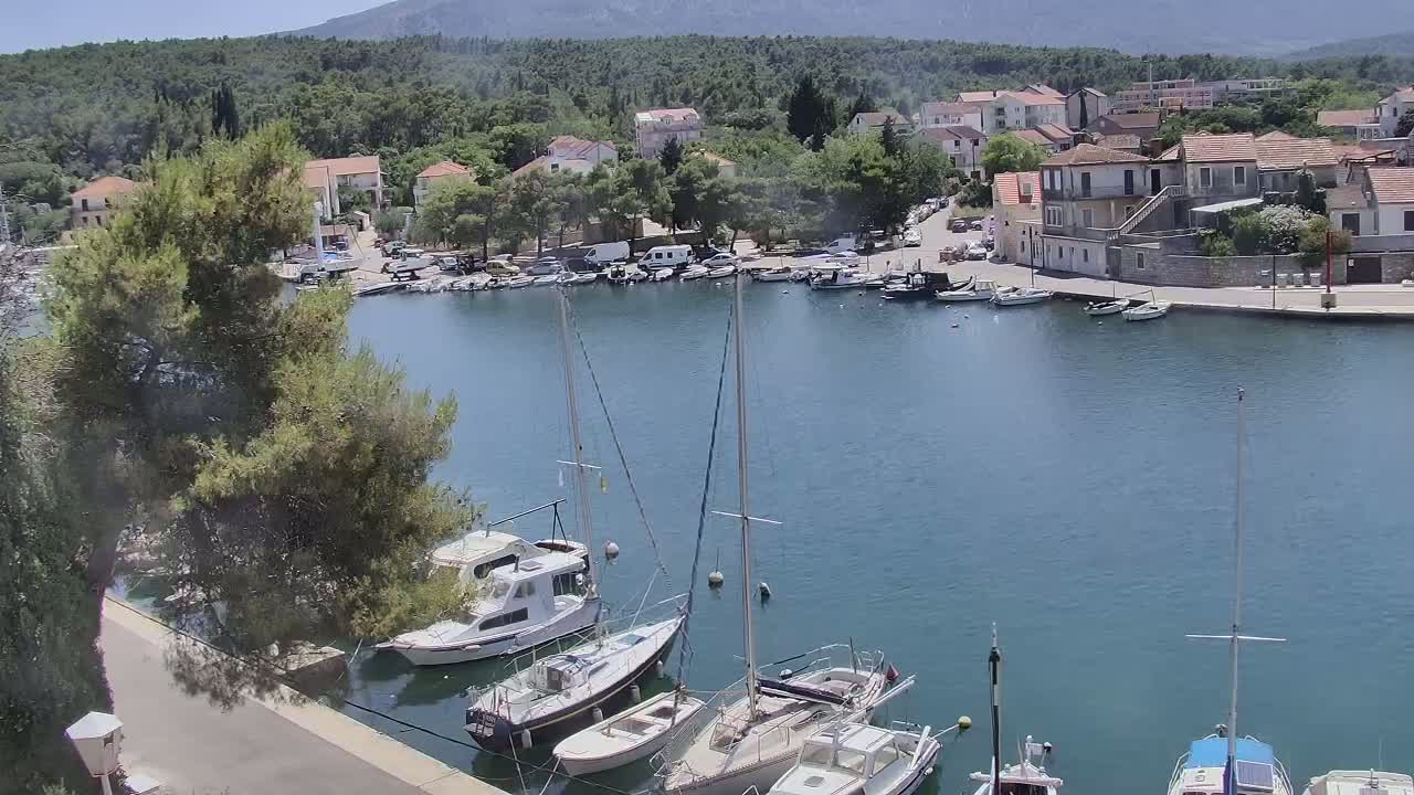 A calm, sunny day reveals a harbor filled with sailboats and motorboats, nestled beside a small town surrounded by green hills and mountains.