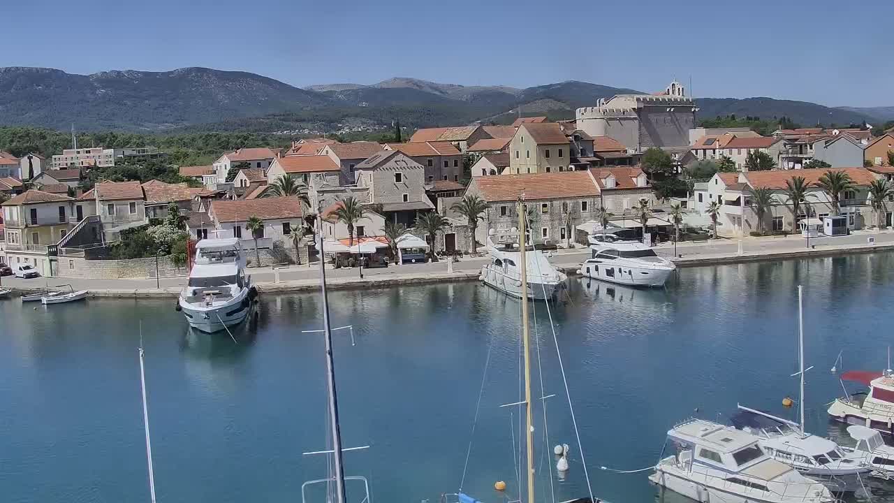 A waterfront town with numerous boats in a calm harbor, backed by low mountains under a clear blue sky.