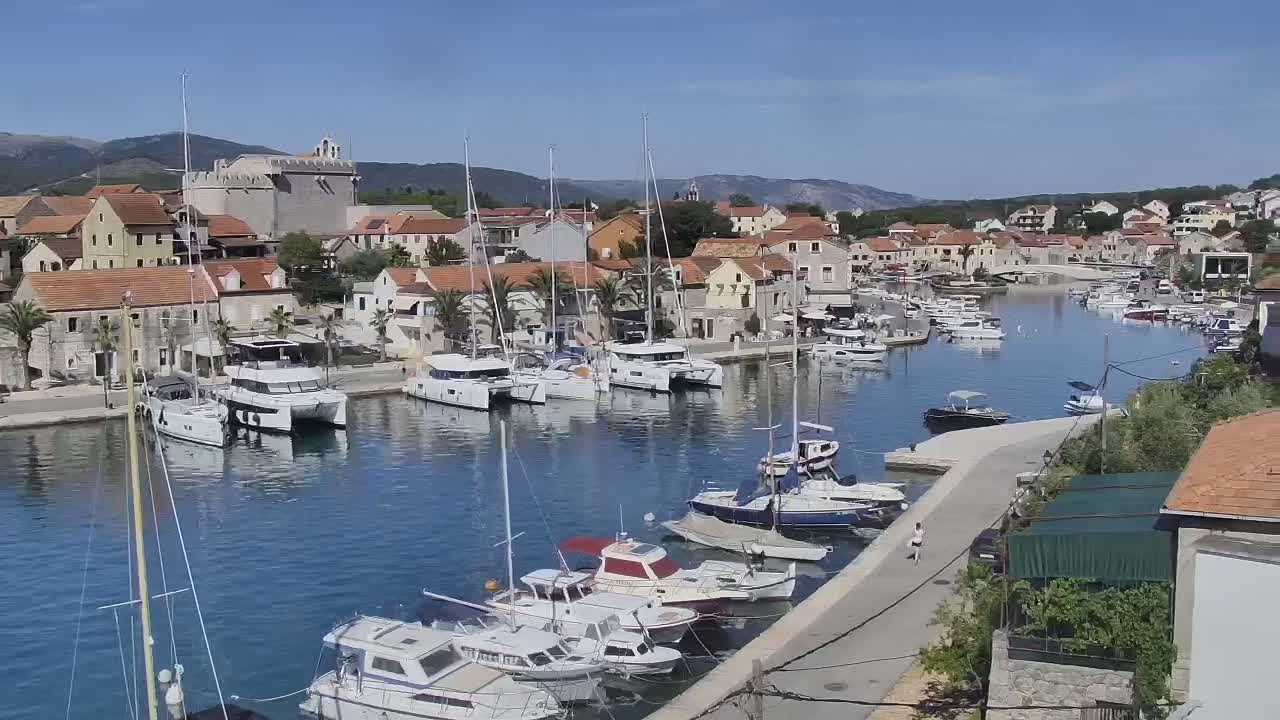 A harbor filled with sailboats and motorboats is nestled in a town of low stone buildings under a clear blue sky.