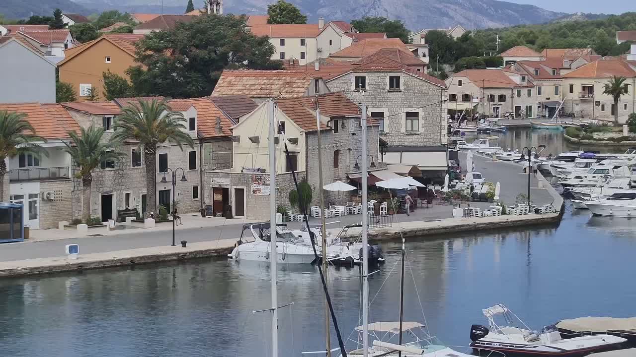 A waterfront town with stone buildings, palm trees, and numerous boats in a calm harbor, set against a backdrop of mountains on a clear day.