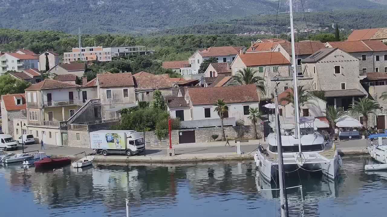 A waterfront village of low, red-roofed buildings with a docked catamaran and other boats, all under a sunny sky.