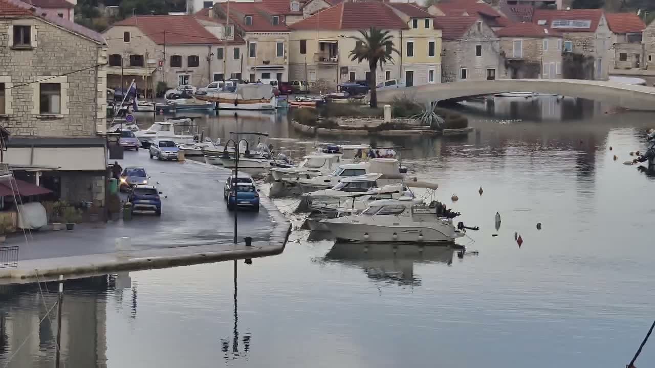 A tranquil harbor scene on an overcast day shows numerous boats docked along a wet, car-lined road, with traditional stone buildings and a bridge framing the background.