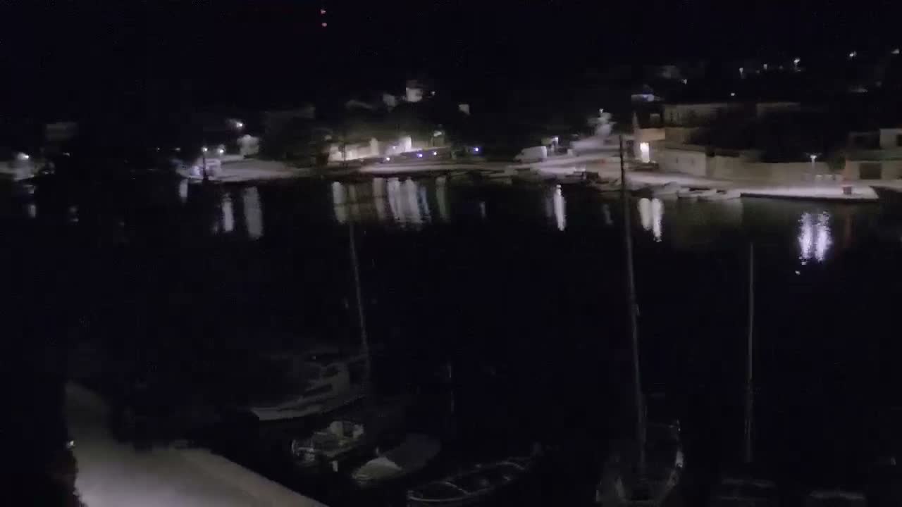 A serene nighttime view captures a coastal town across calm dark waters reflecting distant lights, with several boats moored in the foreground under a clear sky.