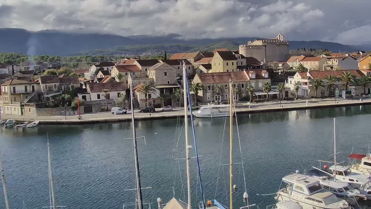 Masts of sailboats rise from a calm harbor in the foreground, fronting a coastal town filled with red-roofed buildings and palm trees lining a waterfront road, with a historic stone fort and distant mountains under a partly cloudy sky.