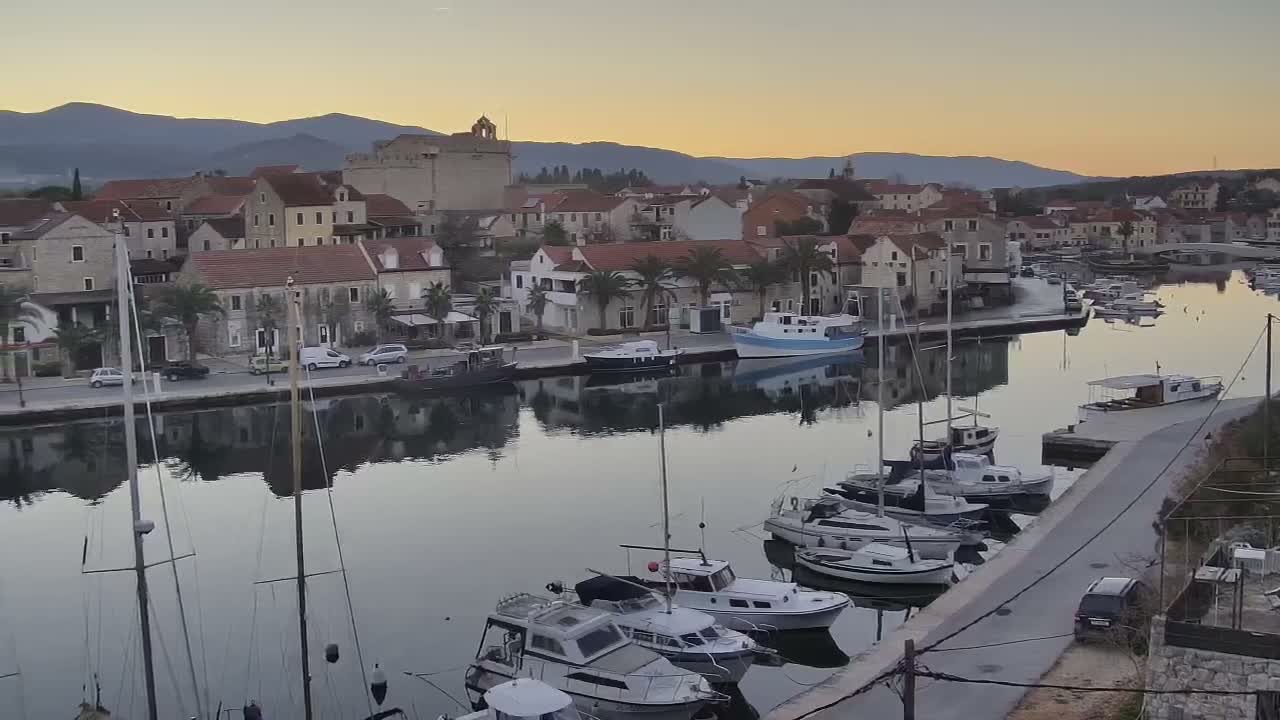 Masts of sailboats rise from a calm harbor in the foreground, fronting a coastal town filled with red-roofed buildings and palm trees lining a waterfront road, with a historic stone fort and distant mountains under a partly cloudy sky.