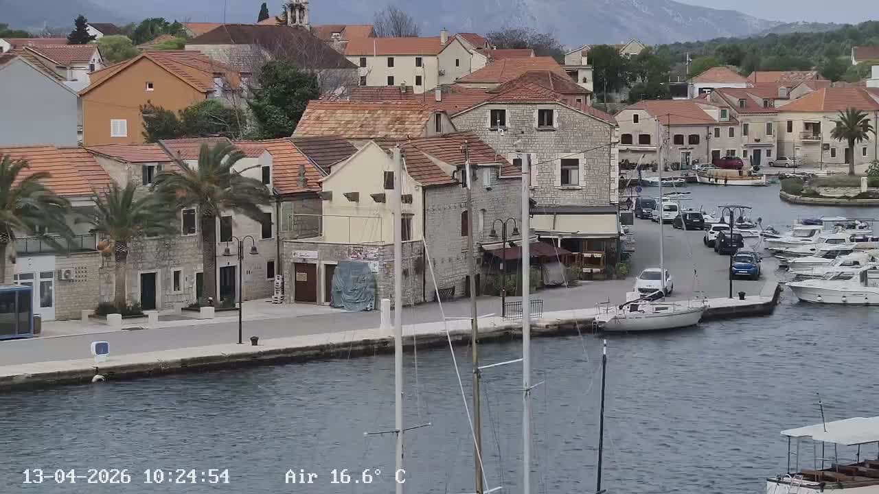 Masts of sailboats rise from a calm harbor in the foreground, fronting a coastal town filled with red-roofed buildings and palm trees lining a waterfront road, with a historic stone fort and distant mountains under a partly cloudy sky.