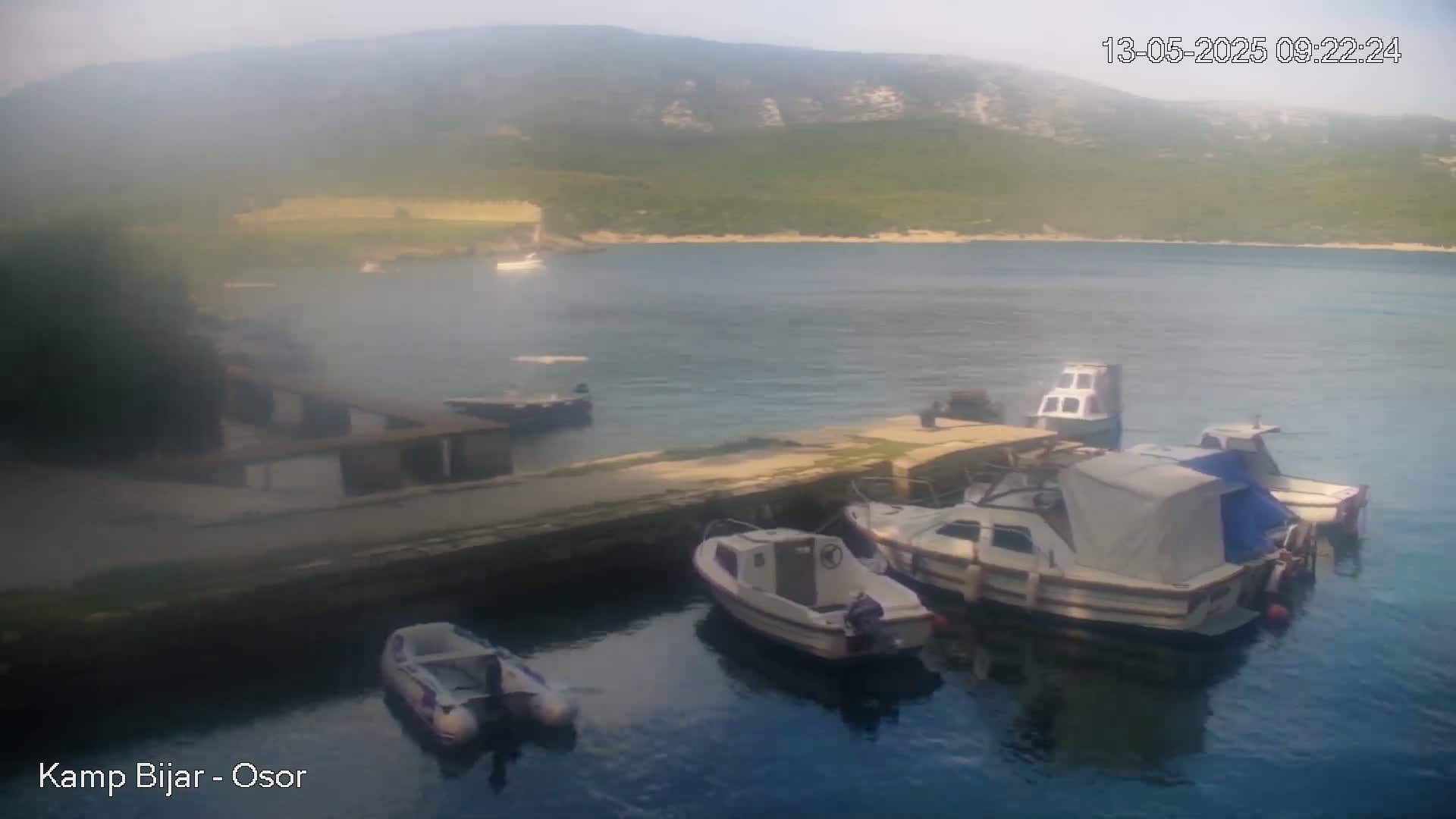 Several small boats are moored at a stone dock next to calm blue water, with hazy green hills visible in the background on a hazy day.