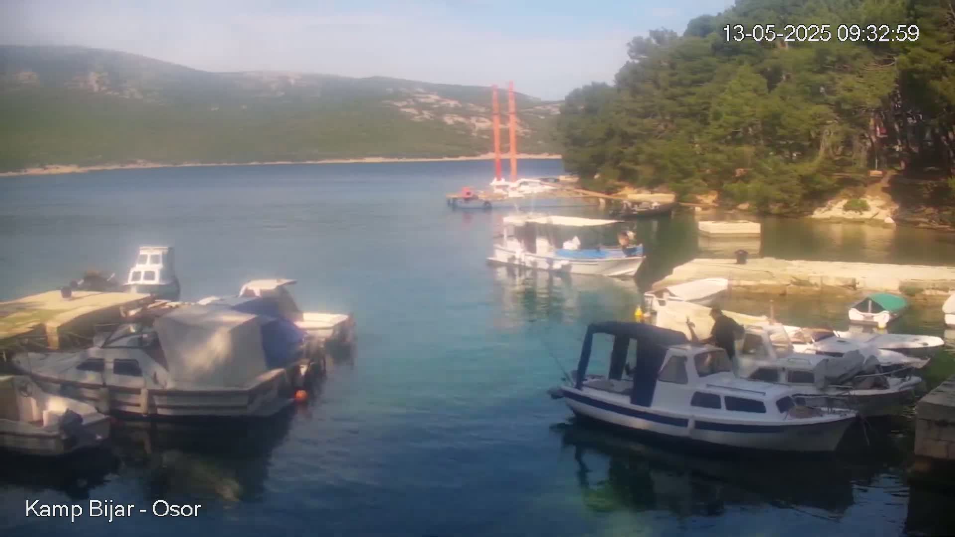 Several boats are moored in a calm, clear blue bay on a sunny day, with green hills and trees in the background.