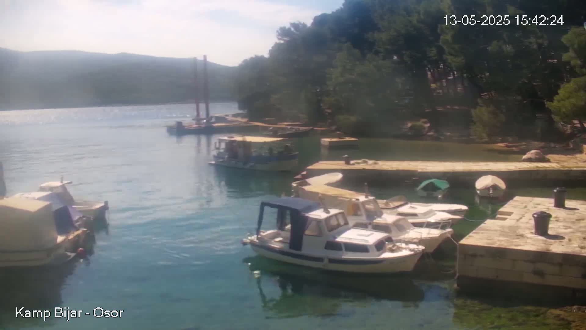 Several small boats are moored in a calm, clear harbor next to a stone jetty, with green hills and trees in the background under a partly sunny sky.