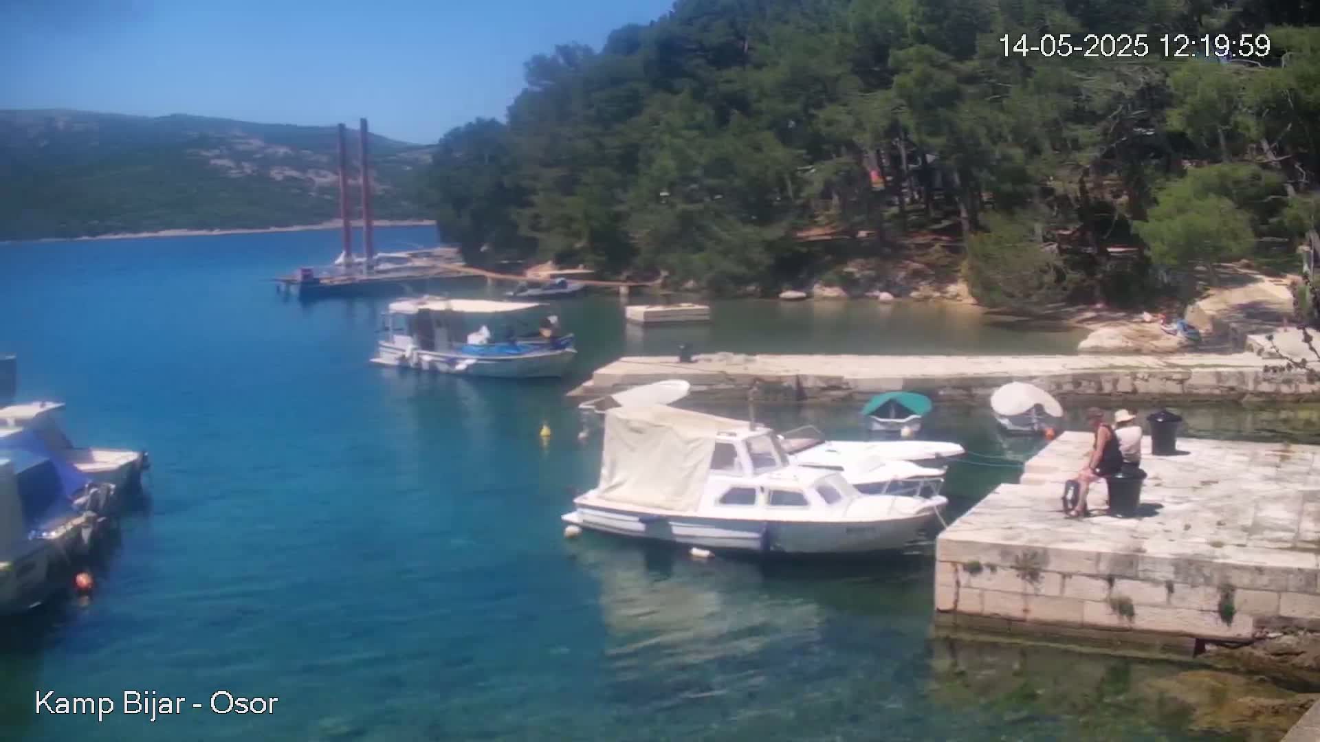 Several boats are docked in a calm, clear blue body of water next to a stone pier, with green hills and trees in the background under a sunny sky.