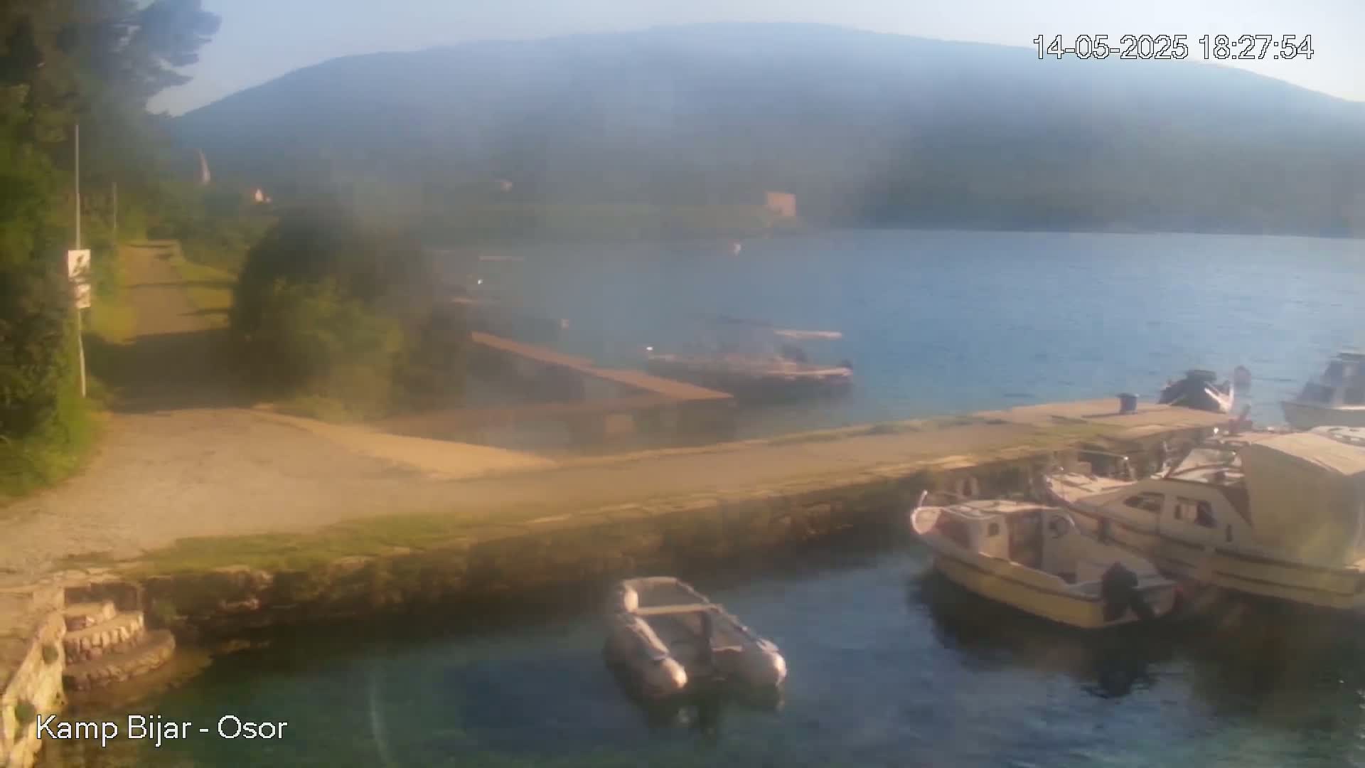 Several small boats are moored at a dock next to a calm body of water, with hazy hills visible in the background under a clear sky.