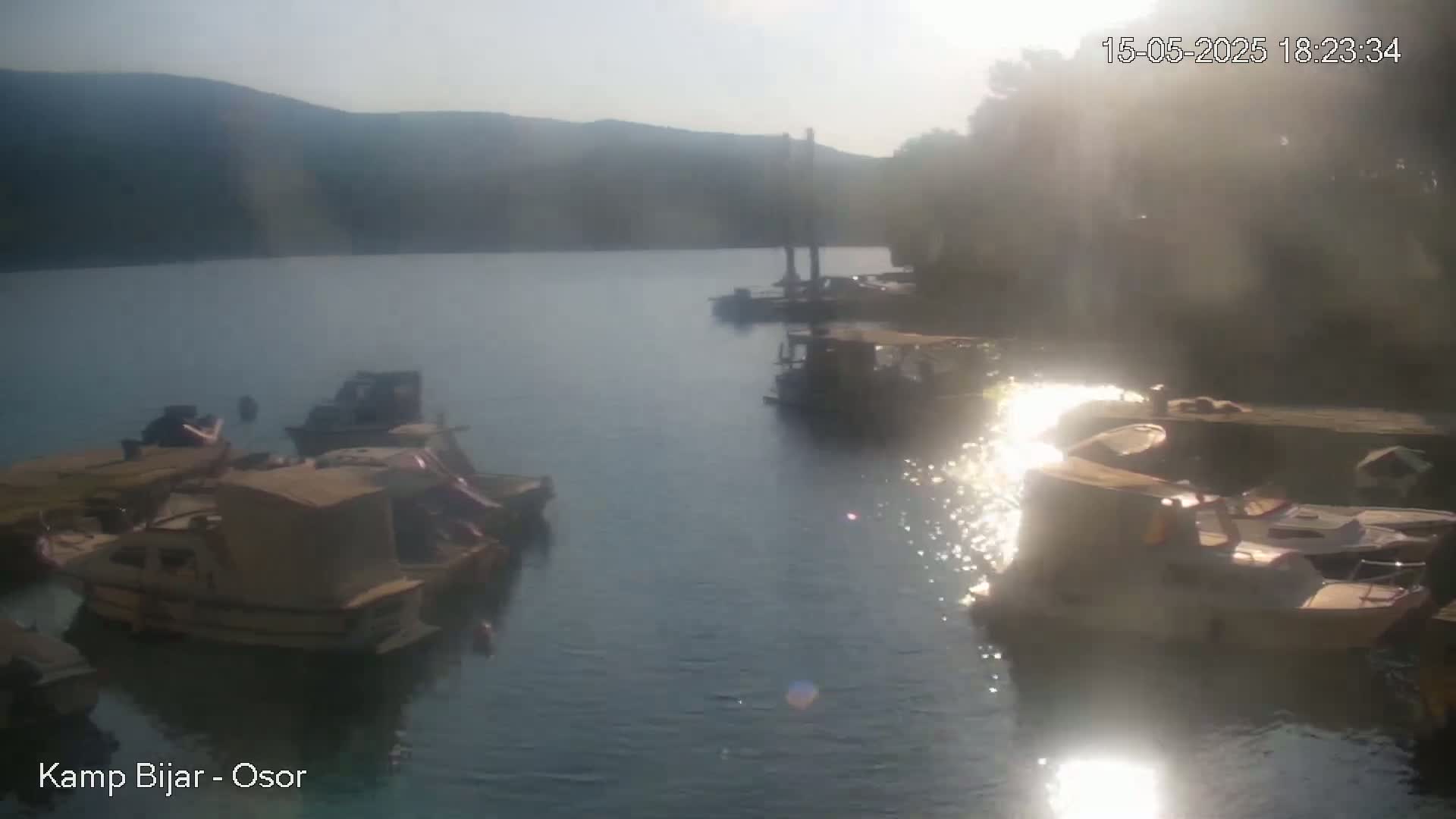 Several boats are moored at a calm, sunlit waterfront, with hazy mountains visible in the background.