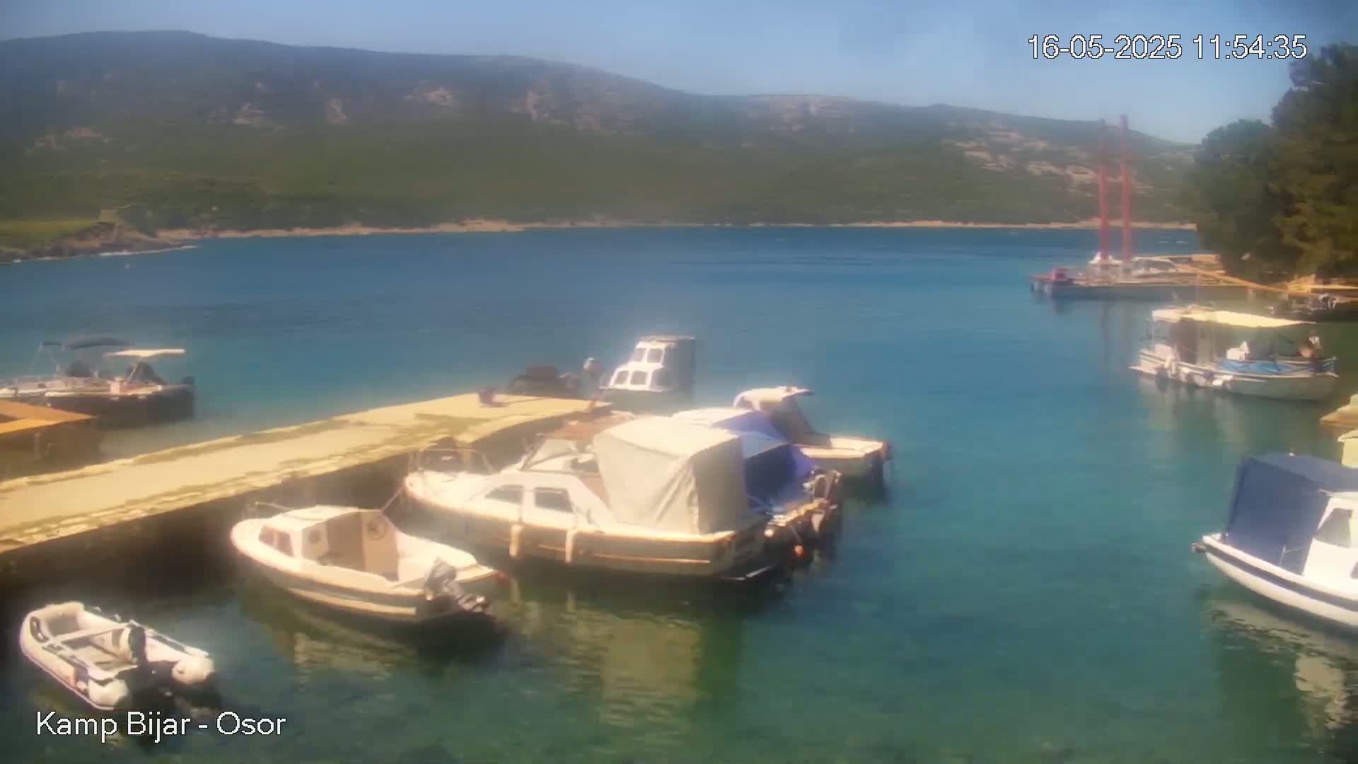 Several motorboats are moored at a calm, clear, teal-colored bay under a sunny sky, with green hills in the background.