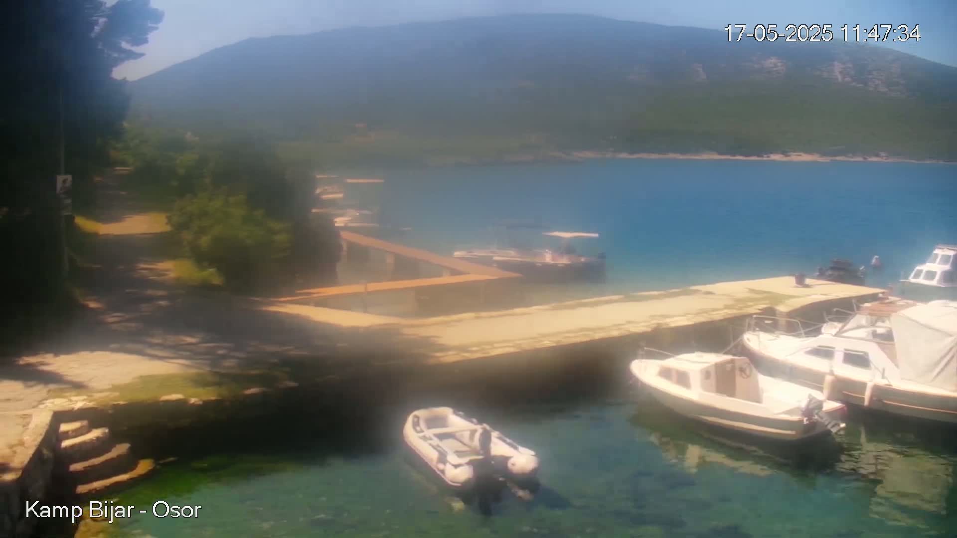 A hazy, sunny day shows a tranquil bay with several small boats moored at a dock near a green hillside.