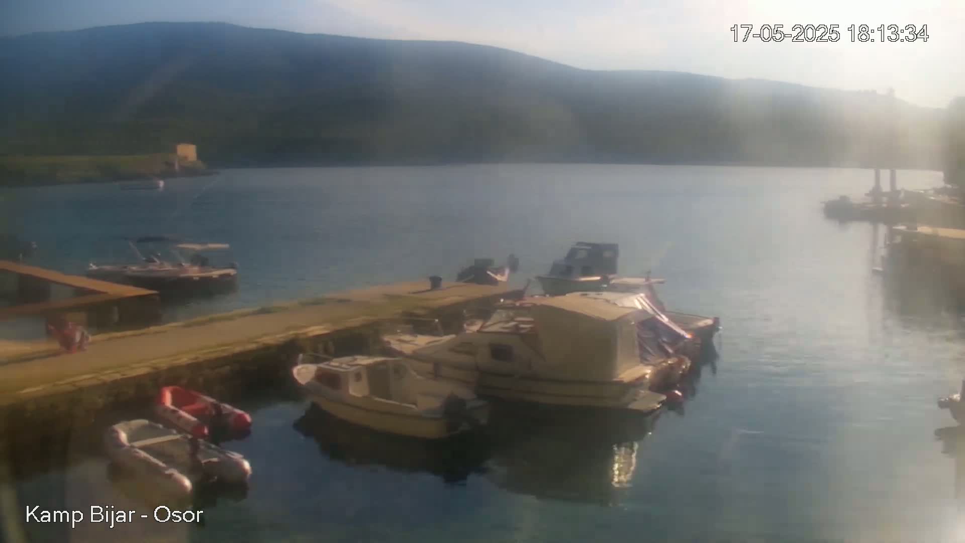 Several boats are docked in a calm harbor, with a hazy sun and distant mountains visible in the background.