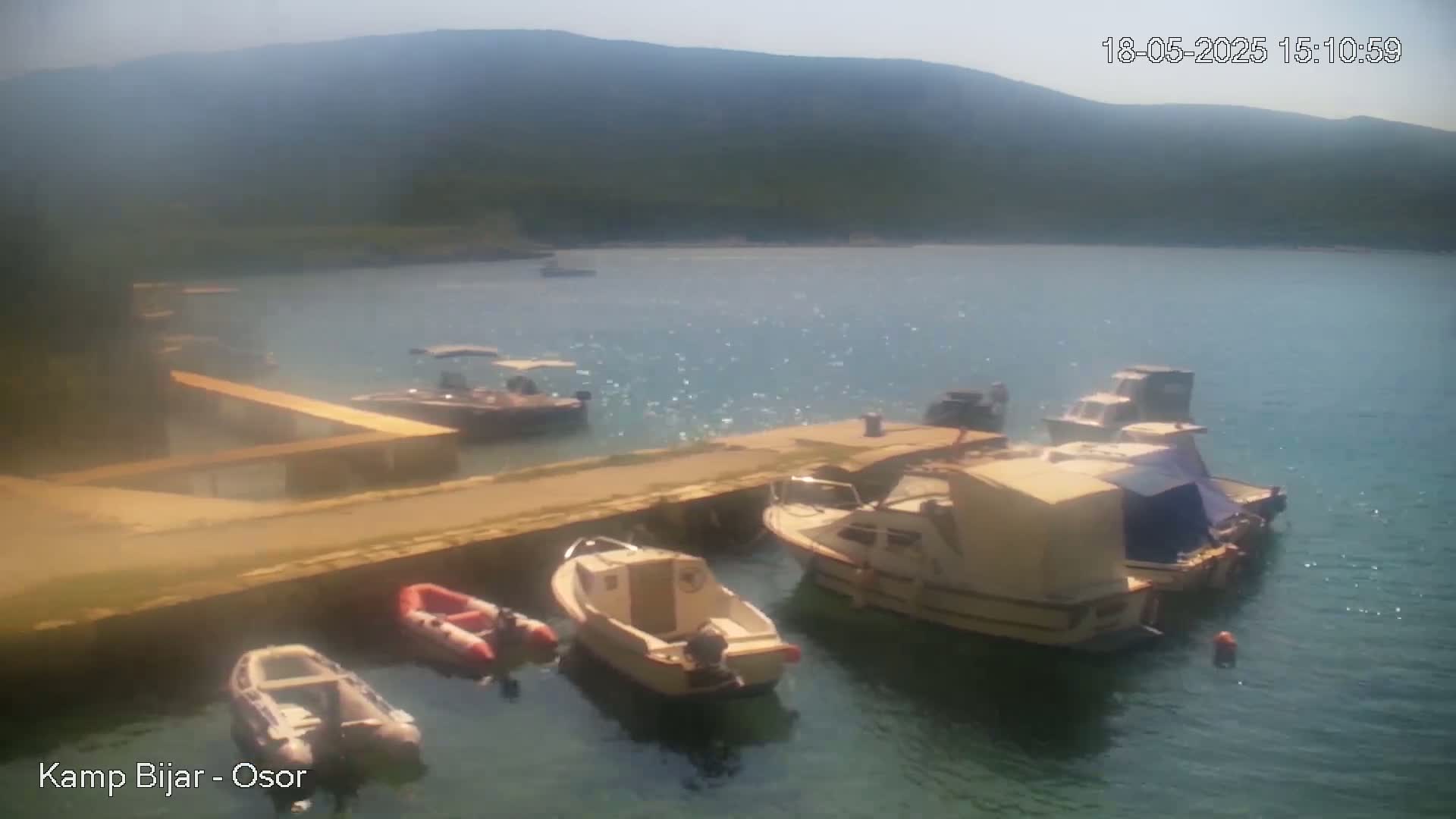 Several boats are moored at a concrete dock in a calm, sunny bay, with mountains visible in the background.