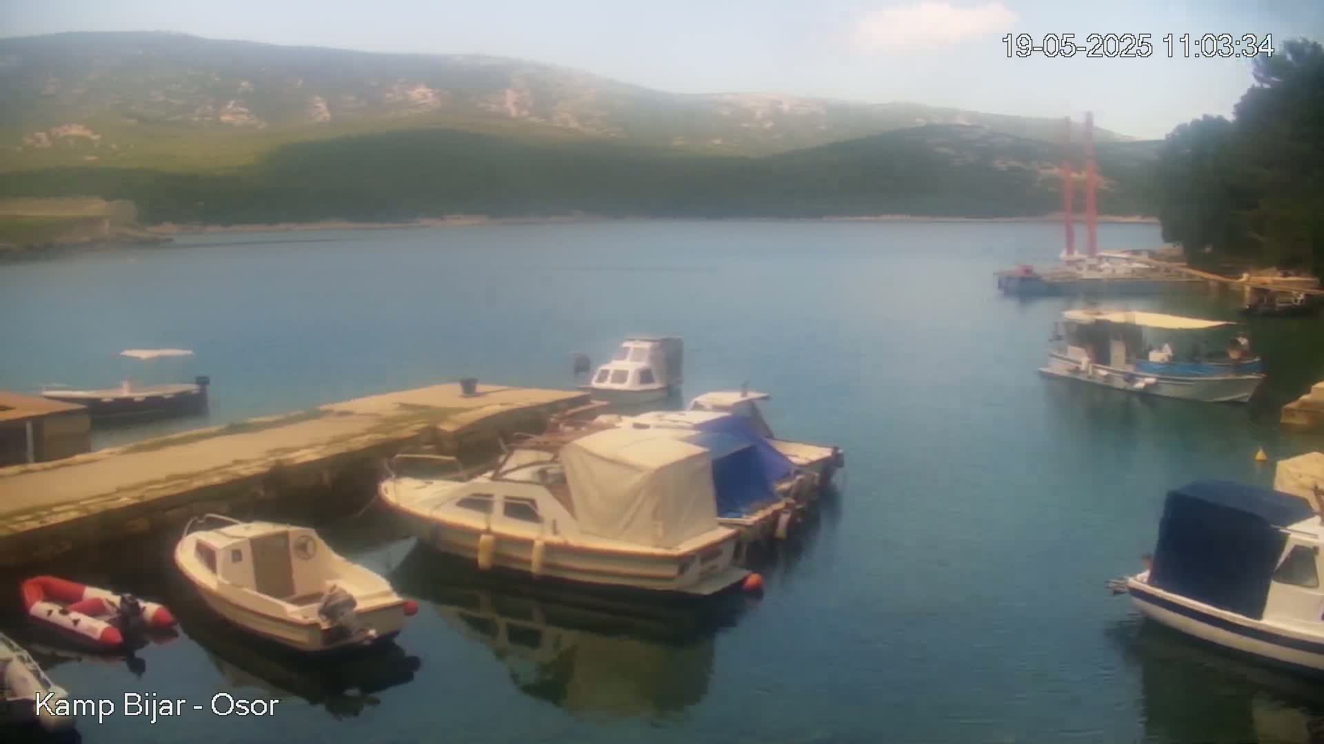 Several boats are moored in a calm bay on a sunny day, with green hills visible in the background.