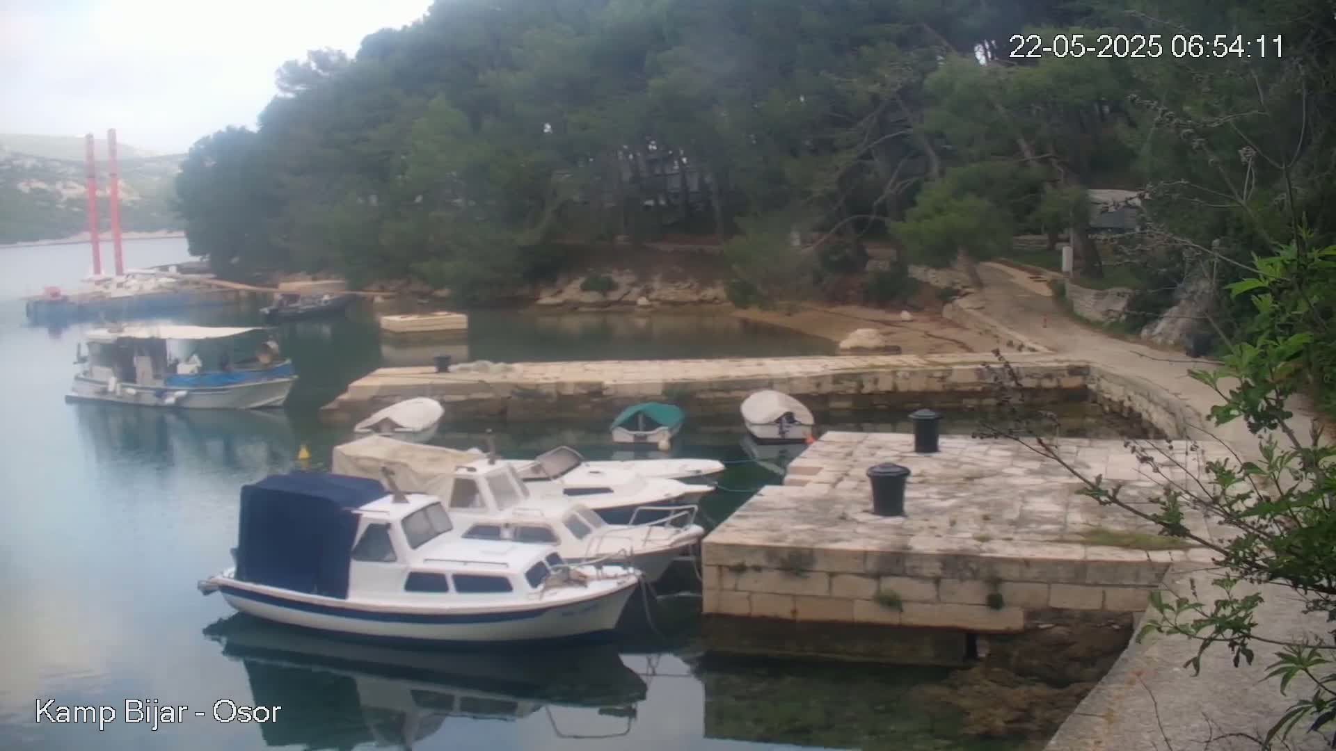 Several small motorboats are moored at a stone dock in a calm bay surrounded by lush green trees under an overcast sky.