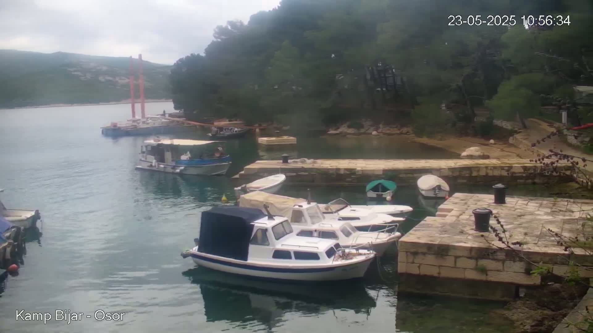 Several small motorboats are moored at a stone dock in a calm bay under an overcast sky, with a forested hillside in the background.