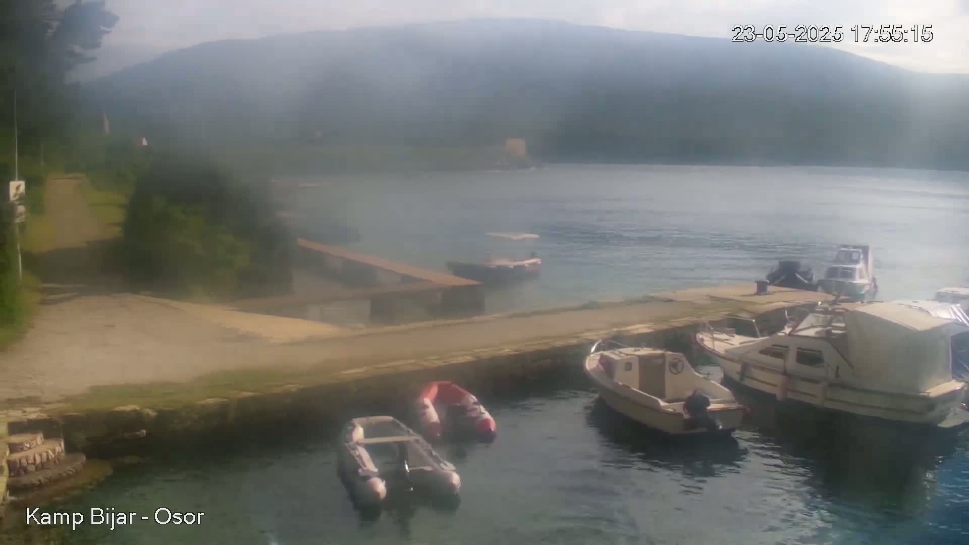 Several motorboats and small inflatable boats are moored at a dock in a calm bay, with hazy hills visible in the background under a partly cloudy sky.