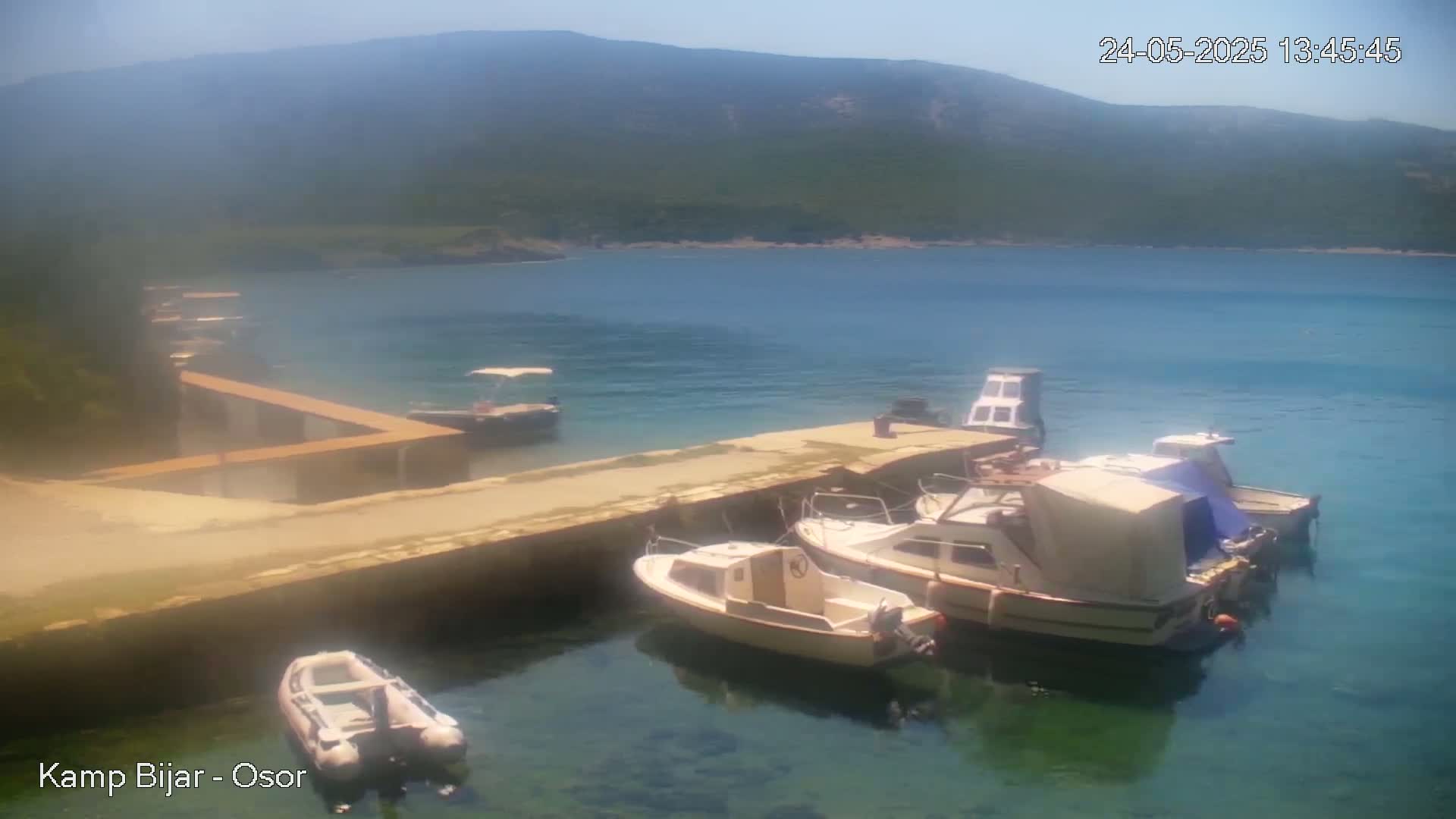 Several small boats are moored at a concrete dock next to a calm, clear body of water under a sunny sky, with a mountain visible in the background.