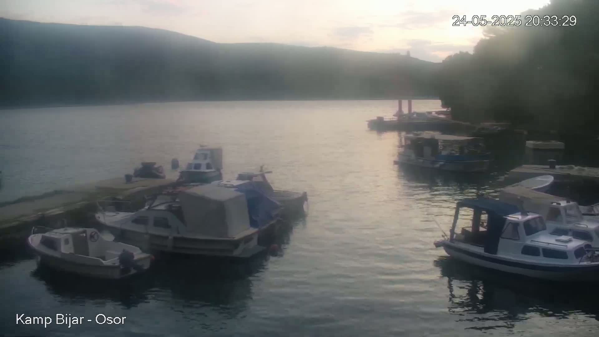 Several small boats are moored at a calm waterfront, with hills visible in the background under a dusky sky.