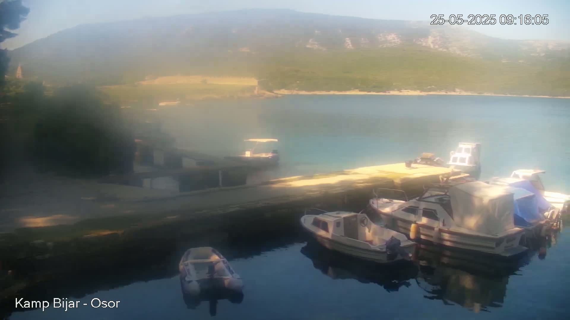 Several small boats are moored at a dock overlooking a calm, hazy body of water and a distant, green-covered hillside under a bright, slightly hazy sky.