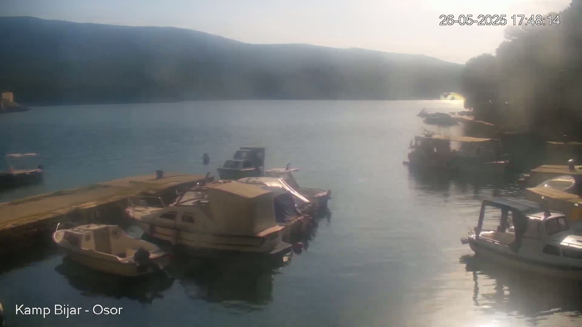 Several boats are moored at a dock on a calm body of water under a hazy, sunny sky, with hills visible in the background.