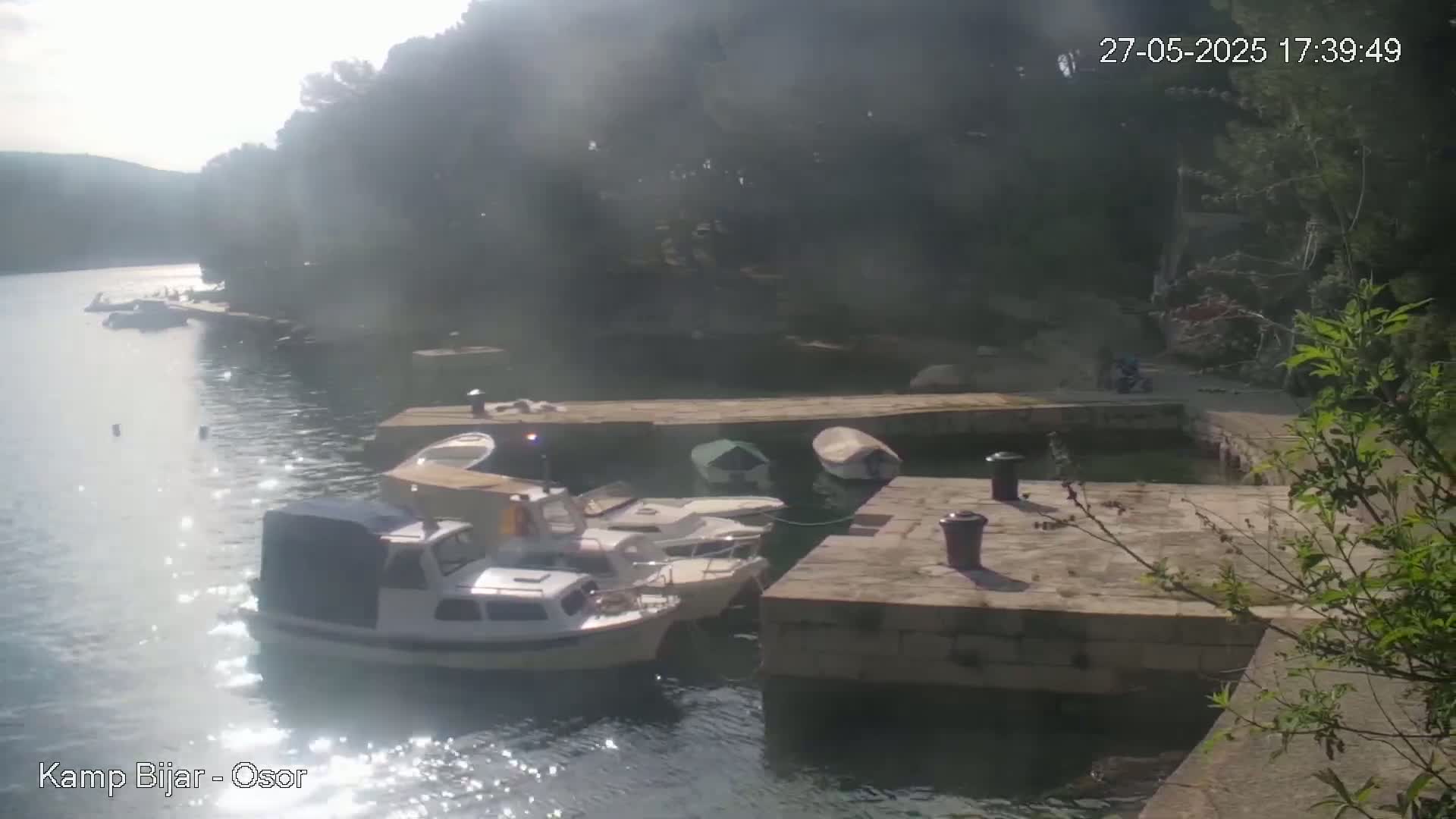 Several small motorboats are docked at a stone pier on a sunny day, next to calm water and lush green vegetation.