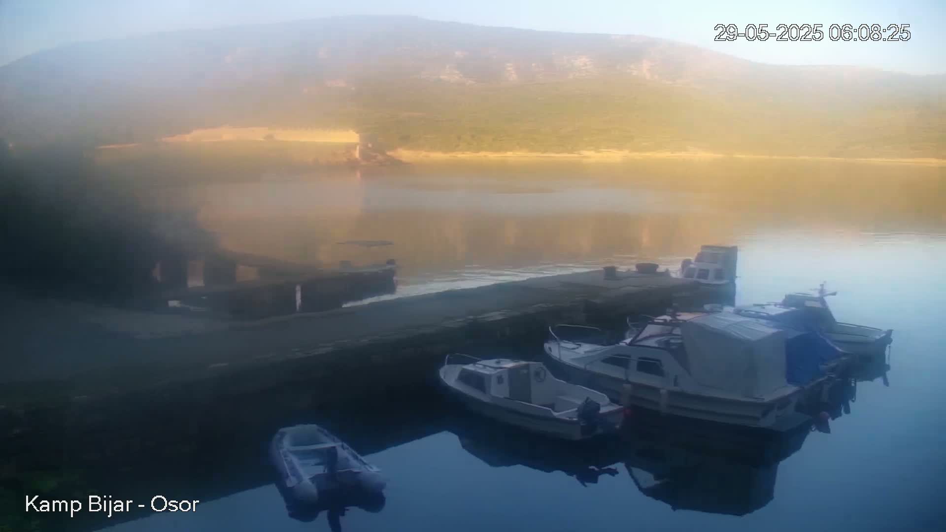 Several small boats are moored at a dock on a calm, misty body of water at sunrise, with hills visible in the background.