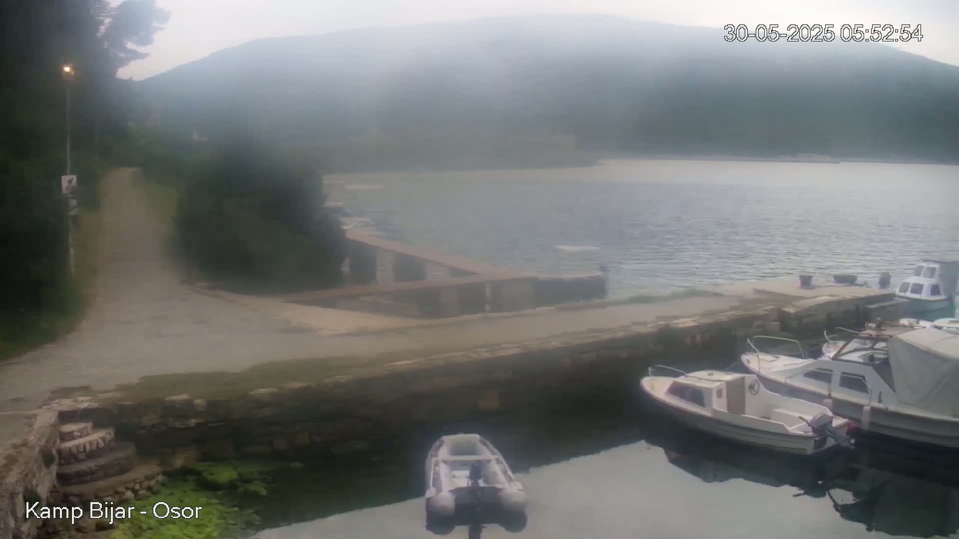 A hazy, overcast day reveals a small harbor with several boats docked, a paved path leading to the water, and a hill shrouded in mist in the background.