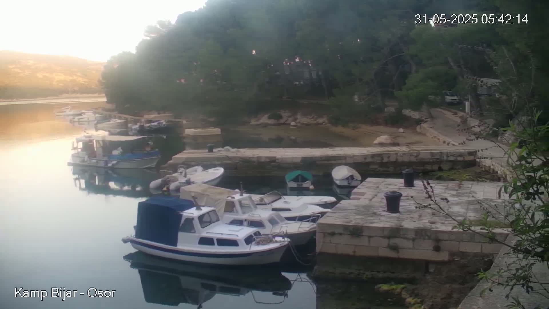 Several small motorboats are moored at a stone dock next to a calm body of water, with hazy sunlight illuminating the scene and lush green vegetation bordering the shore.