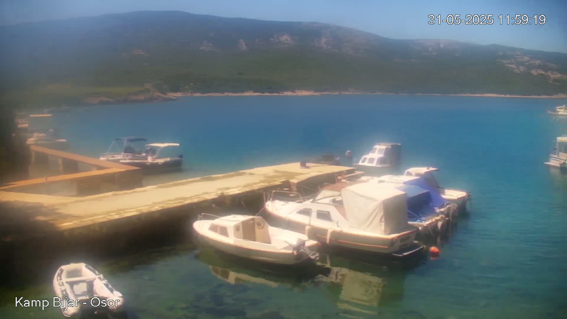 Several small boats are moored at a dock on a calm, clear blue body of water, with mountains visible in the background under a sunny sky.