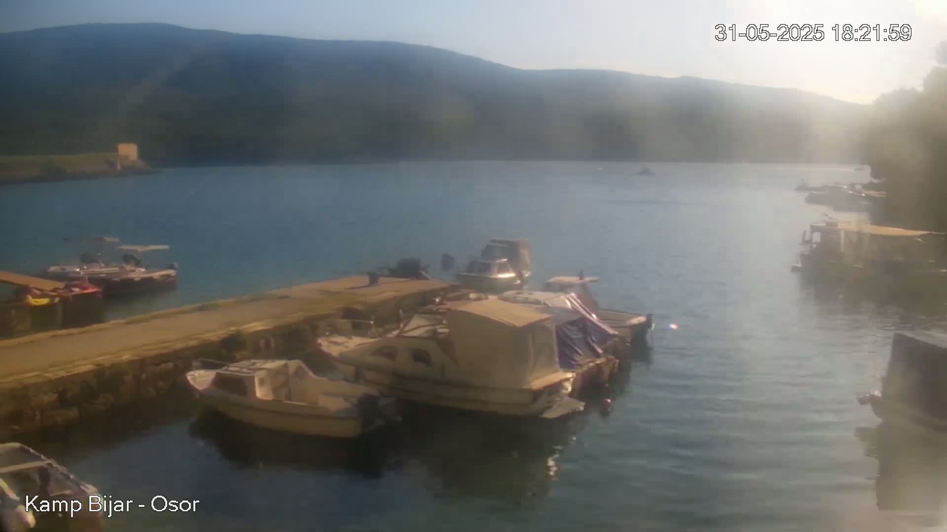 Several boats are moored at a waterfront dock on a calm, sunny day, with mountains visible in the background.