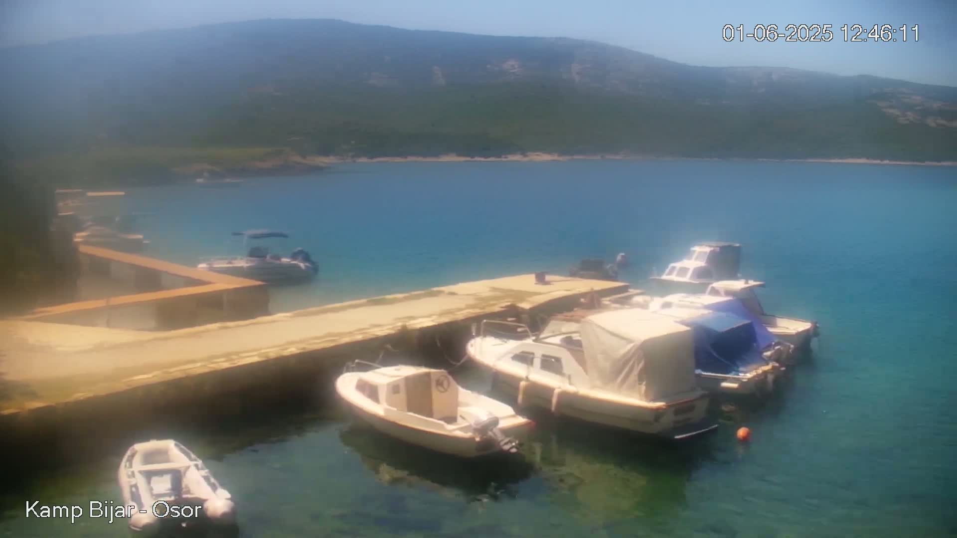 Several small motorboats are moored at a concrete dock in a calm, clear bay under a sunny sky, with mountains visible in the background.