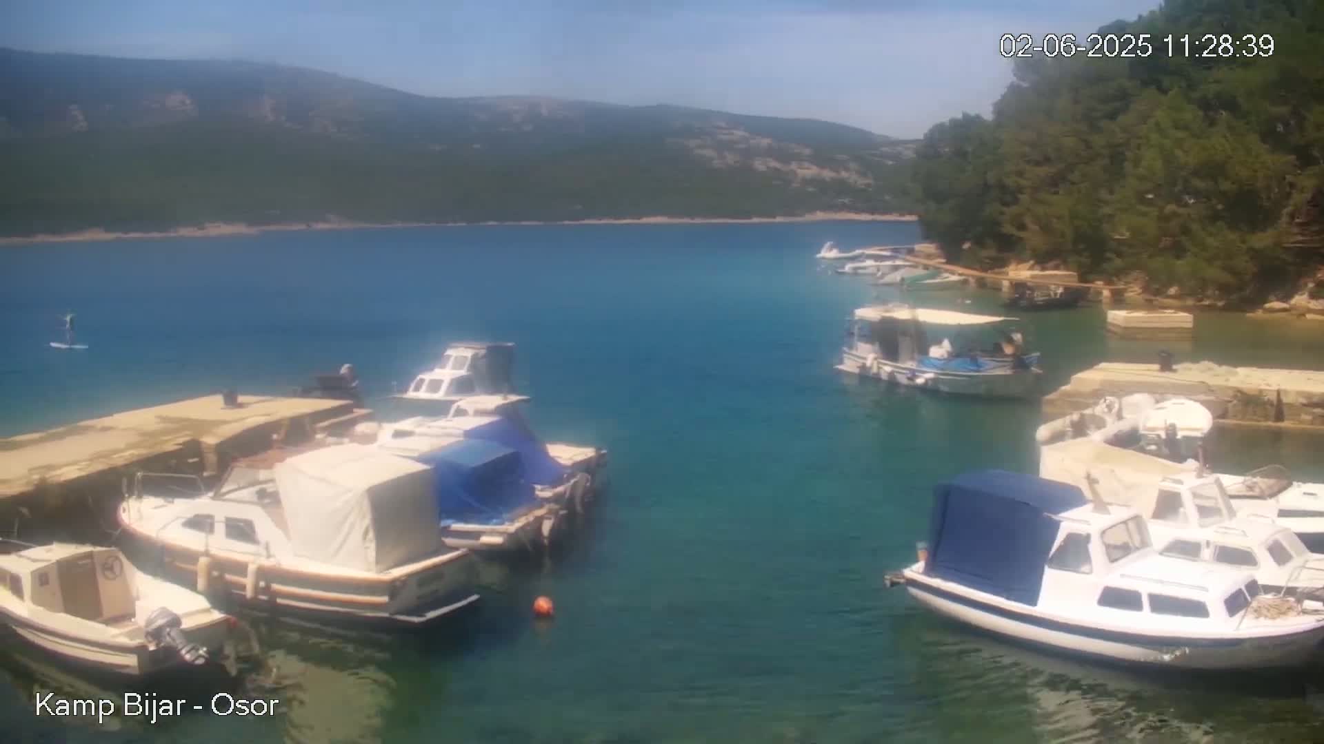 Several boats are moored in a calm, clear blue bay, surrounded by green hills under a sunny sky.