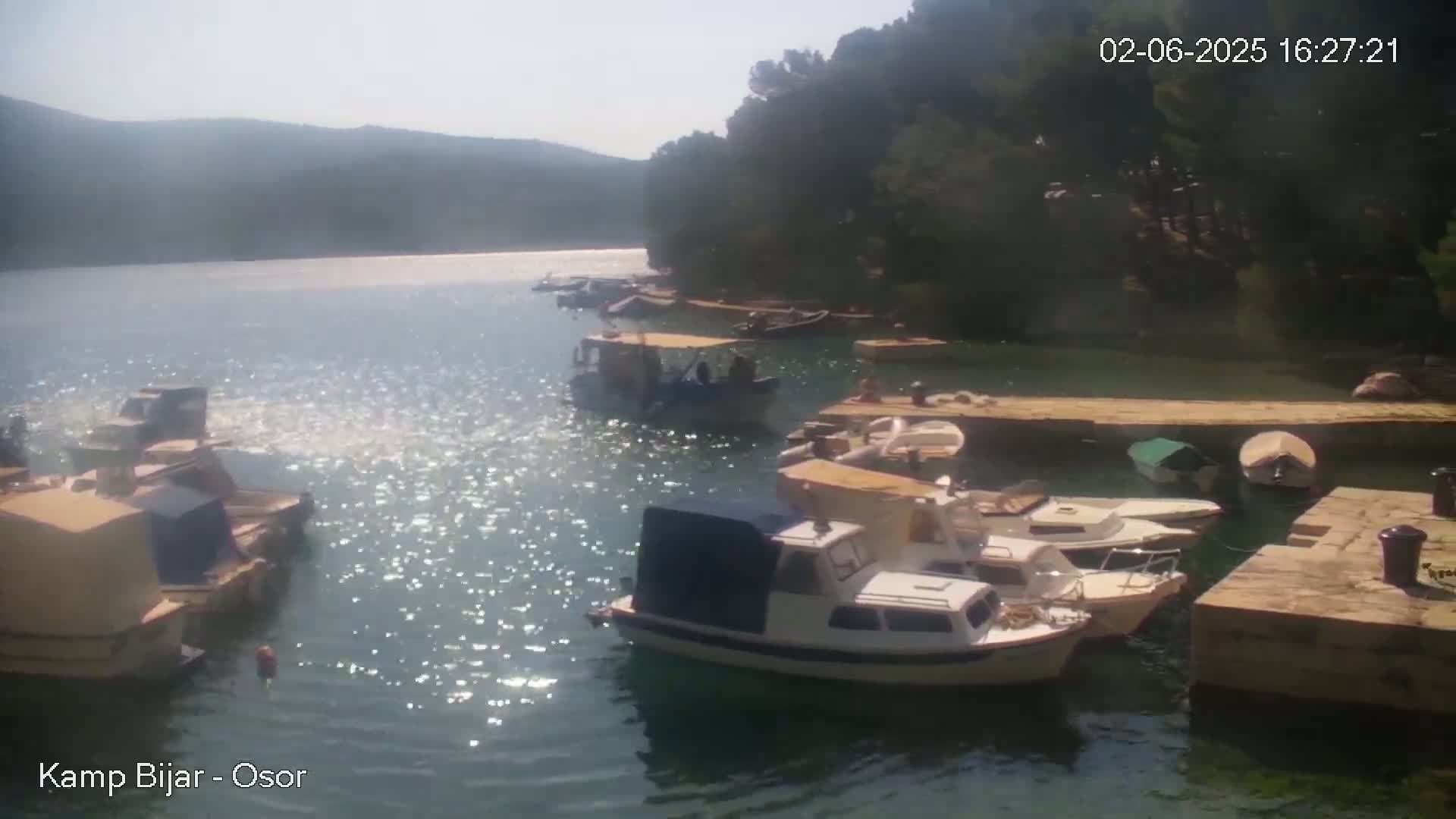 Many small boats are moored in a calm, sunny bay next to a stone dock, surrounded by green hills and trees.