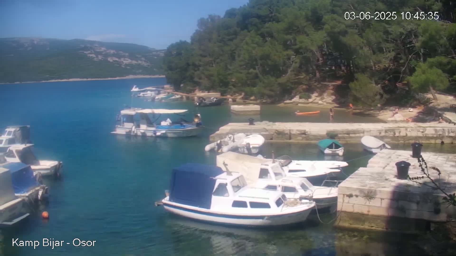 Several boats are moored in a calm, clear blue bay on a sunny day, surrounded by lush green vegetation and a rocky shoreline.