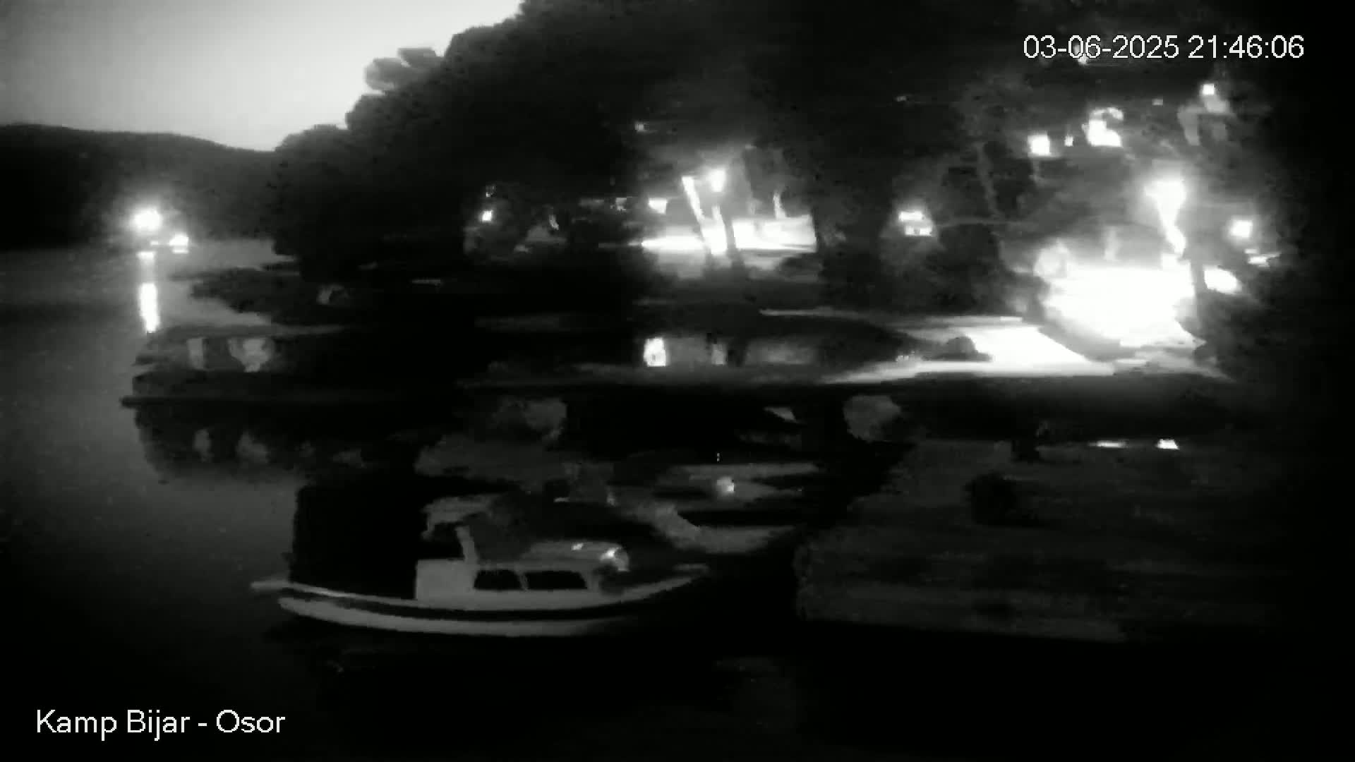 A nighttime, grayscale image shows several vehicles parked near a body of water under a dark sky.