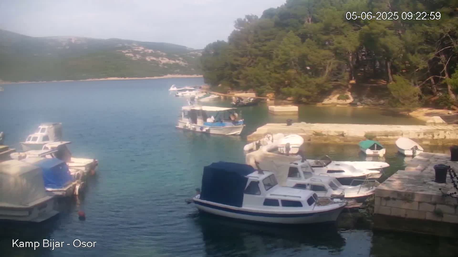 Several boats are moored in a calm bay on a sunny day, surrounded by green hills and trees.