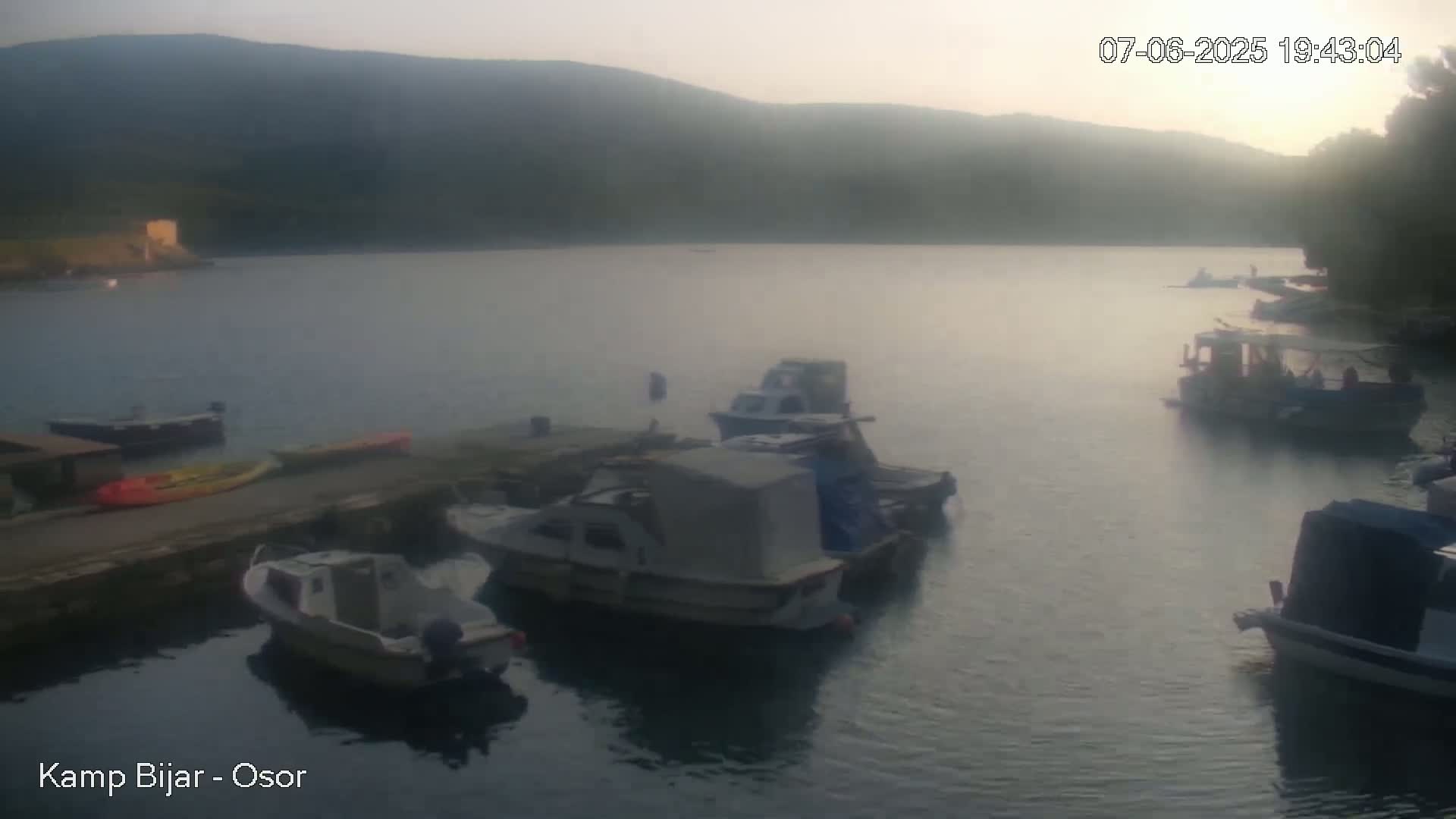 Several small boats are moored at a calm waterfront at dusk, with hills visible in the background.