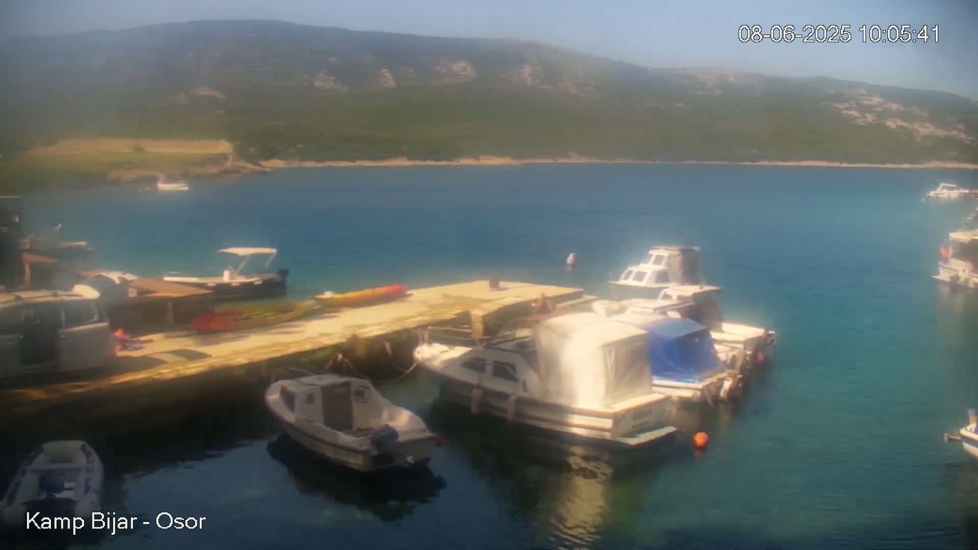 Several boats are docked at a pier in a calm, clear blue bay, with a mountainous, green landscape visible in the background under a sunny sky.