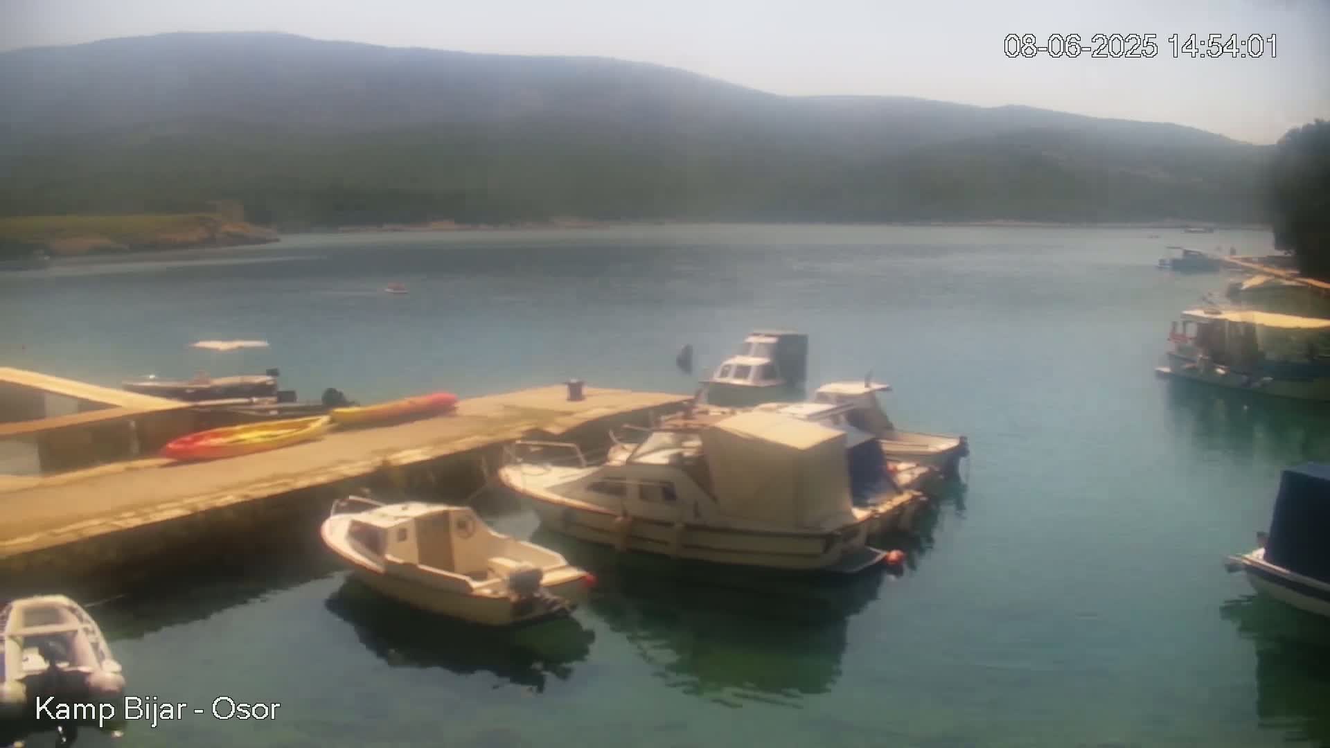 Several boats are moored at a dock in a calm bay, under a hazy sky, with hills in the background.