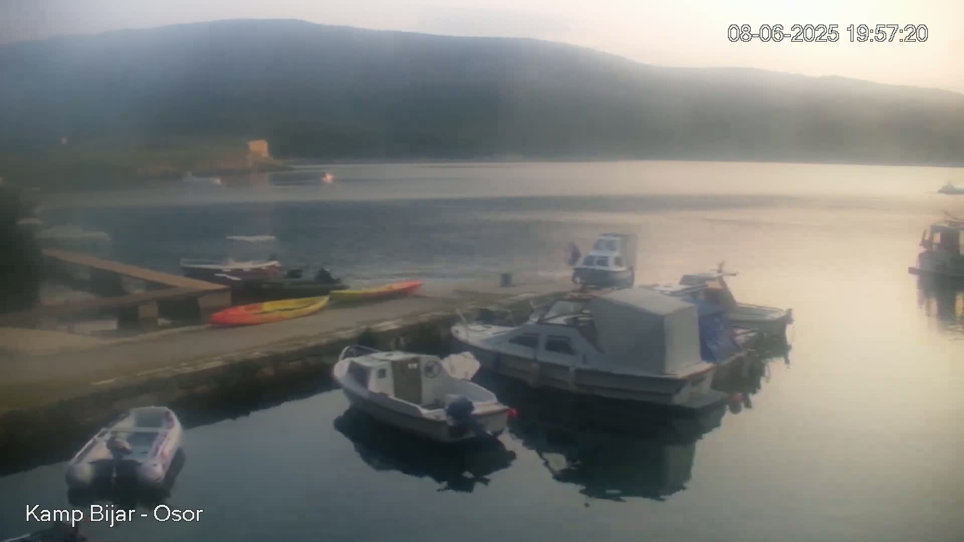 Several boats are moored at a calm harbor, nestled against a hillside under a hazy, late afternoon sky.