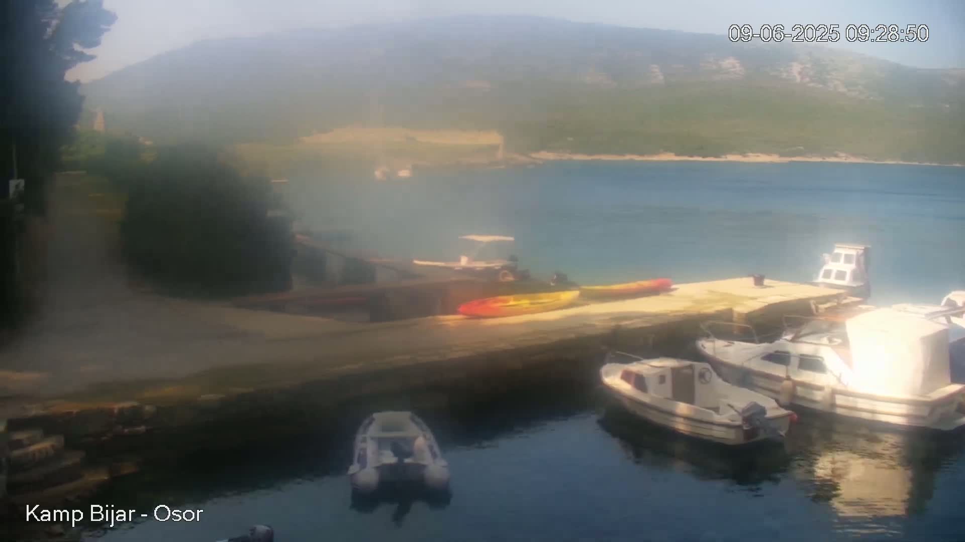 Several small boats are moored at a dock on a calm, hazy day, with a distant view of hills and a body of water.