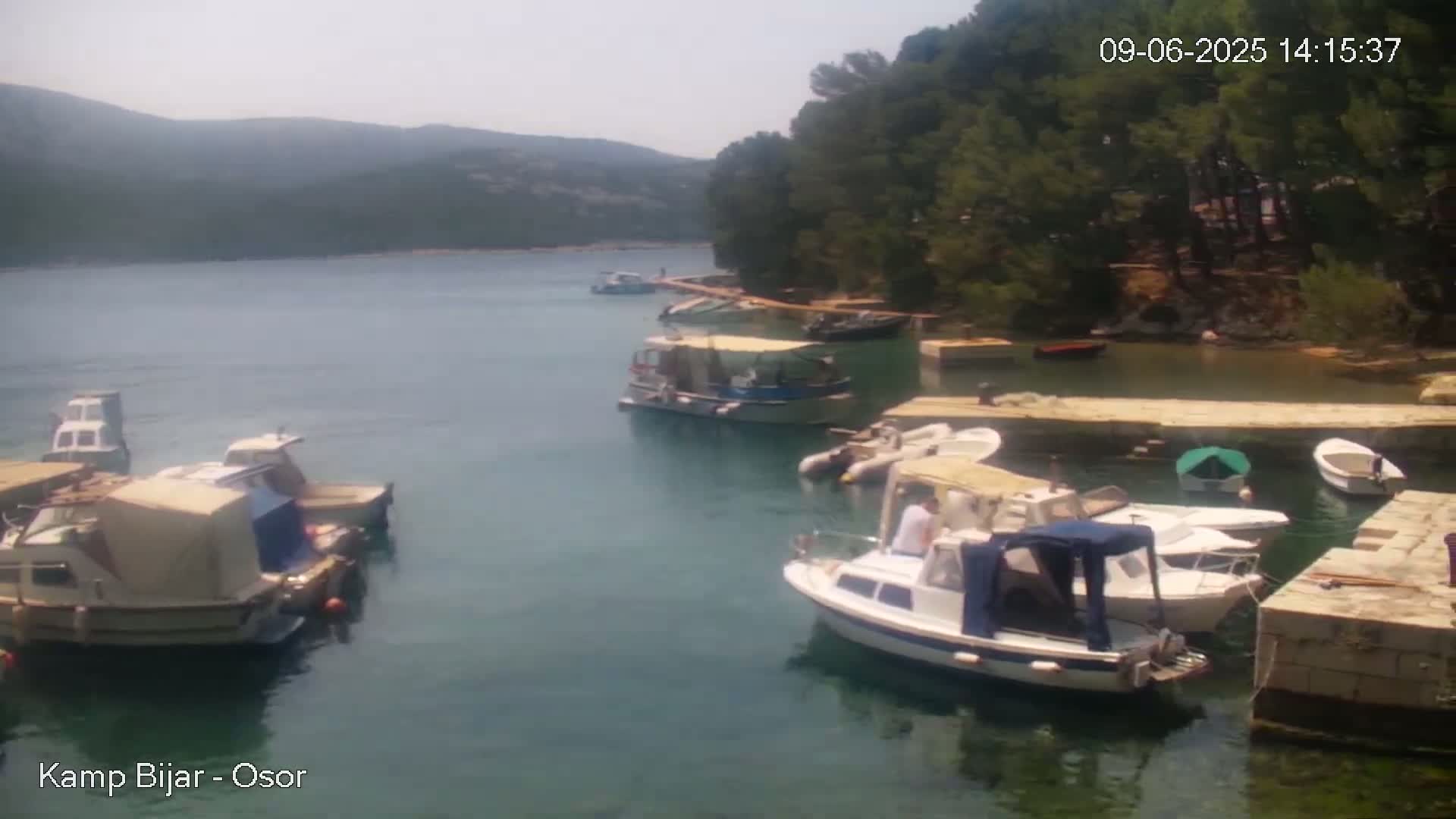 Several small motorboats are moored in a calm, clear bay next to a rocky shore lined with trees under a hazy, sunny sky.
