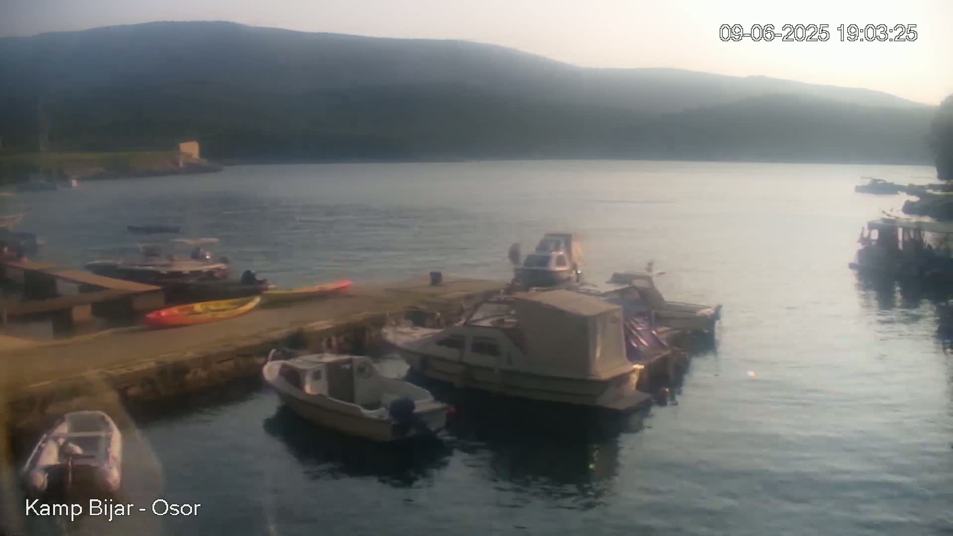 Several boats are docked at a calm waterfront, with kayaks and other small watercraft nearby, under a hazy, light sky against a mountainous backdrop.