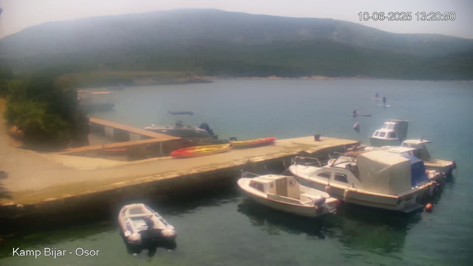 Several motorboats and kayaks are moored at a dock in a calm bay, with hazy mountains in the background and people paddleboarding in the water.